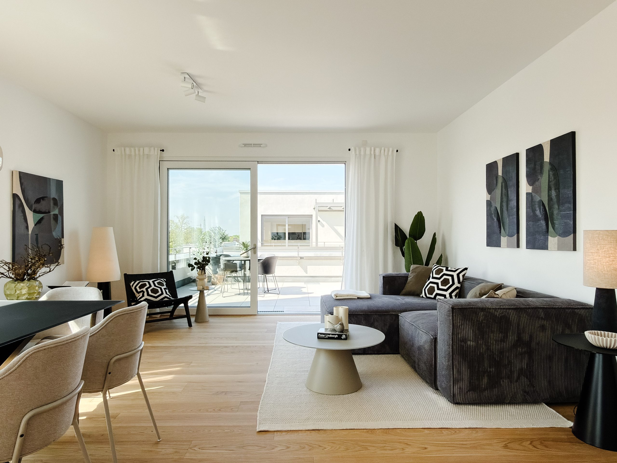 IMG_0103 Bright modern living room with a gray sectional sofa, abstract wall art, plants, and large glass doors to a balcony.